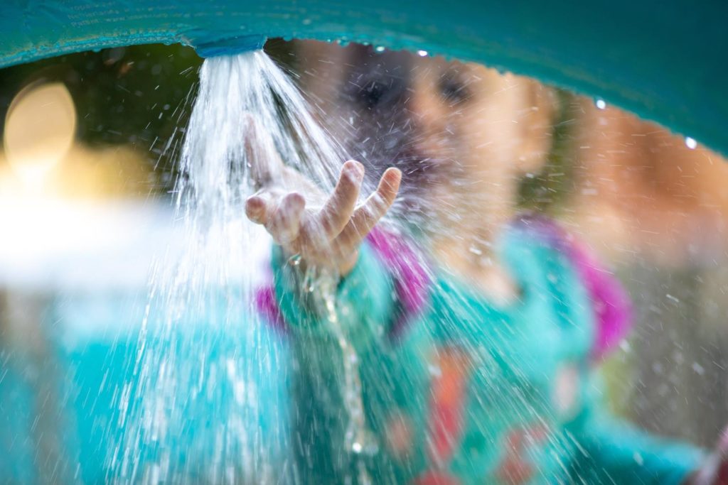 Niña disfrutando de sus colonias de verano: pone su mano bajo el chorro de agua en una de las actividades al aire libre.