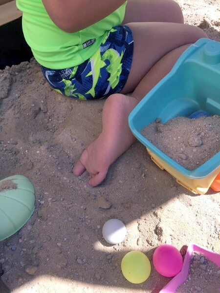 Niño jugando desclazo dentro de un arenero en las colonias de verano de la escuela infantil inclusiva enDARA. Al lado tiene con un cubo con arena dentro y unas pelotas pequeñas de colores