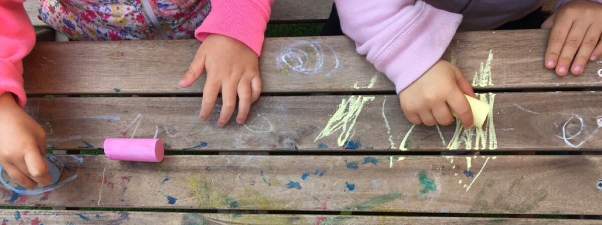 Detalle de las manos de unos niños dibujando con tizas de colores la superficie de una mesa de madera en el patio de la escuela.